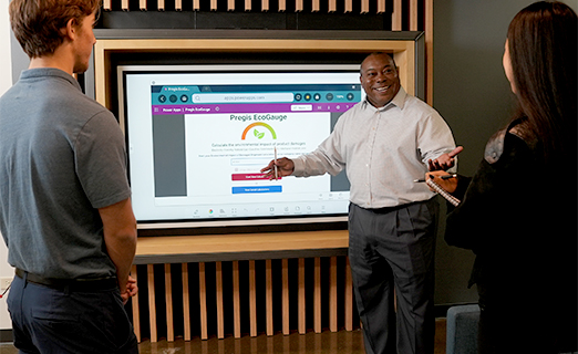 Three professionals in a modern office setting gather around a large digital screen embedded in a wooden frame. One person presents a webpage titled "Pregis EcoGauge," which features a green leaf logo and buttons labeled "Start Assessment" and "Try it now!" The page focuses on calculating the environmental impact of product packaging, suggesting a discussion on sustainability and eco-friendly practices.