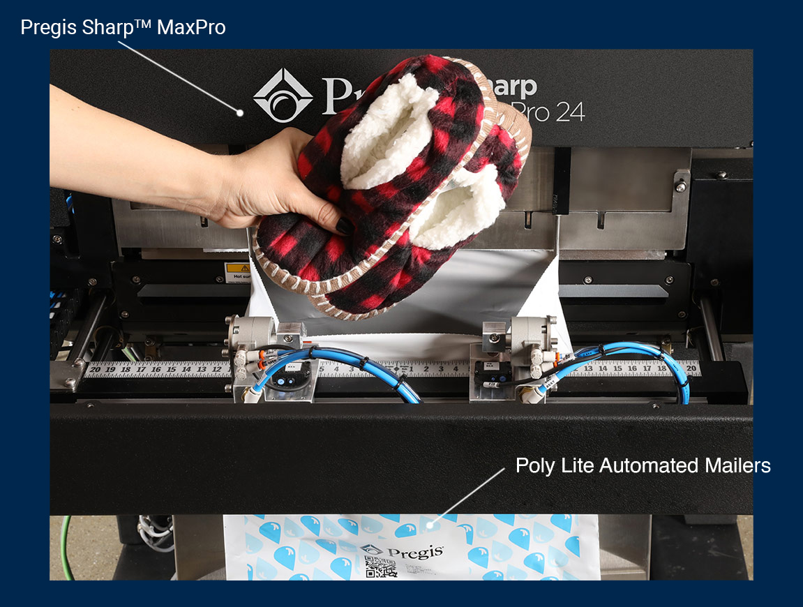 A close-up of the Pregis Sharp™ MaxPro 24 automated packaging system in use. A hand places a red-and-black plaid slipper with white fleece lining into the machine, which is preparing to insert it into a Poly Lite Automated Mailer.